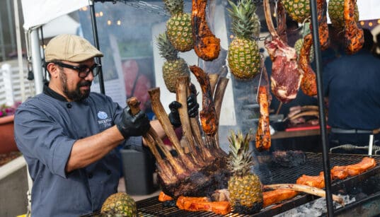 Chef preparing a large rack of meat alongside grilled cuts and pineapples at a previous RARE Steak Championship.