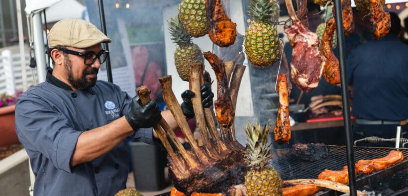 Chef preparing a large rack of meat alongside grilled cuts and pineapples at a previous RARE Steak Championship.