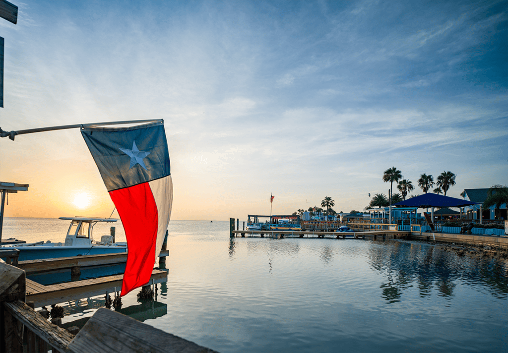 Texas flag overlooking Laguna Madre Bay at sunset on South Padre Island with boats, docks and palm trees in the distance.