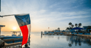 Texas flag overlooking Laguna Madre Bay at sunset on South Padre Island with boats, docks and palm trees in the distance.