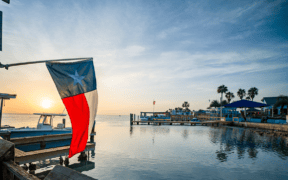 Texas flag overlooking Laguna Madre Bay at sunset on South Padre Island with boats, docks and palm trees in the distance.