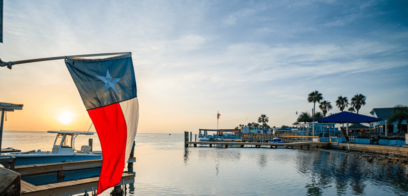 Texas flag overlooking Laguna Madre Bay at sunset on South Padre Island with boats, docks and palm trees in the distance.