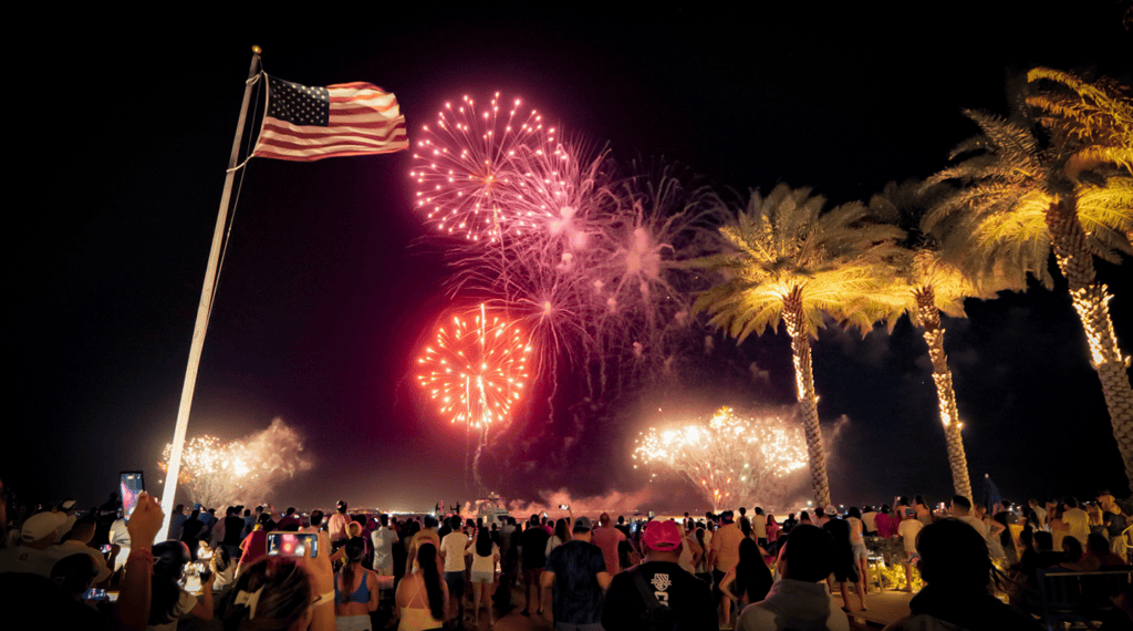 Fireworks burst over South Padre Island as a crowd watches beneath palm trees and an American flag.
