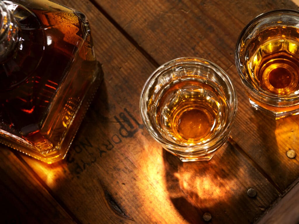 Glasses of amber bourbon beside a bourbon bottle on a rustic wooden table.