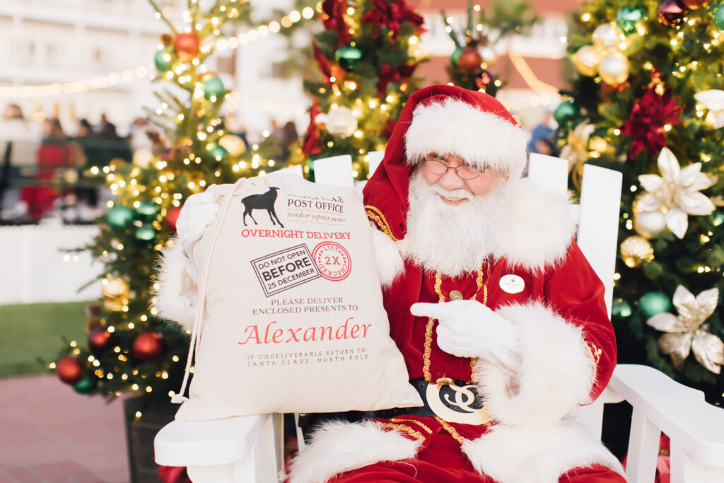 Santa Claus holds a personalized gift sack in front of decorated Christmas trees at Hotel del Coronado.
