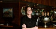 Chef Josh Hood wearing glasses and a black chef’s coat, holding a knife in front of Champagne Tiger’s wood-paneled bar.