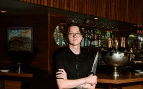 Chef Josh Hood wearing glasses and a black chef’s coat, holding a knife in front of Champagne Tiger’s wood-paneled bar.