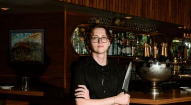 Chef Josh Hood wearing glasses and a black chef’s coat, holding a knife in front of Champagne Tiger’s wood-paneled bar.