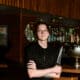 Chef Josh Hood wearing glasses and a black chef’s coat, holding a knife in front of Champagne Tiger’s wood-paneled bar.