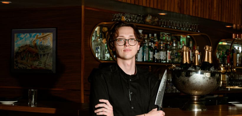 Chef Josh Hood wearing glasses and a black chef’s coat, holding a knife in front of Champagne Tiger’s wood-paneled bar.