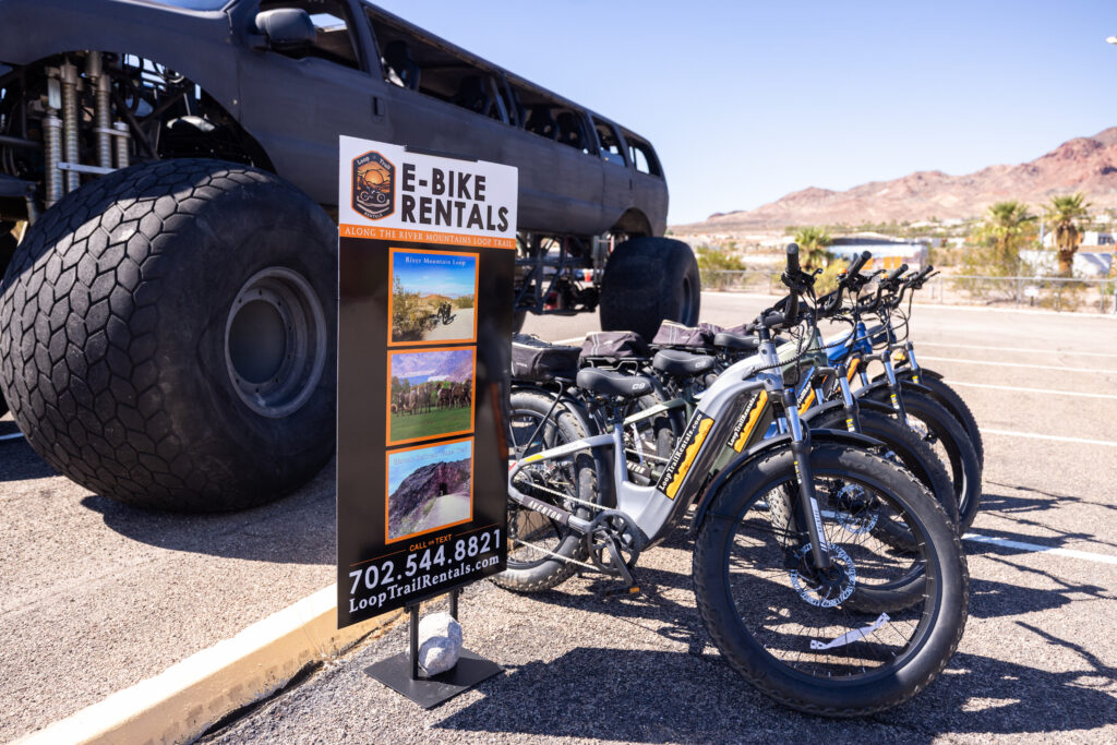 Row of Loop Trail Rentals e-bikes beside a rental sign, with desert mountains in the background at the Boulder City Adventure Center.