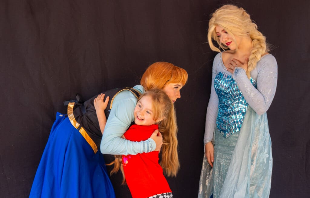 Young girl in a red shirt hugs a costumed princess while another princess in a sparkling blue gown looks on.