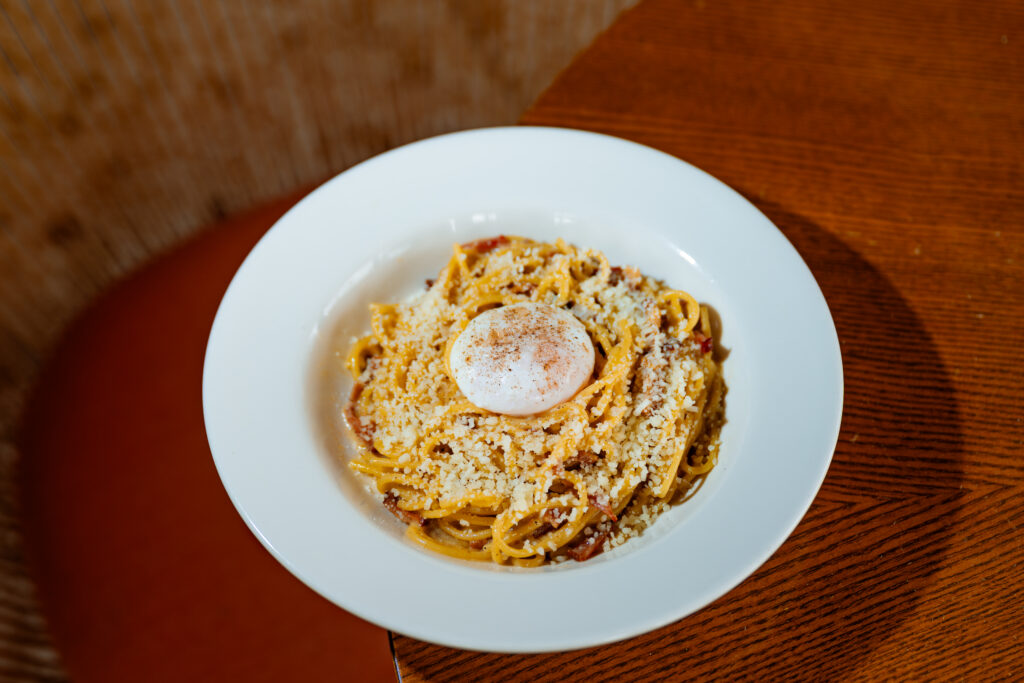 Carbonara breakfast pasta topped with grated cheese and a poached egg in a wide white bowl.