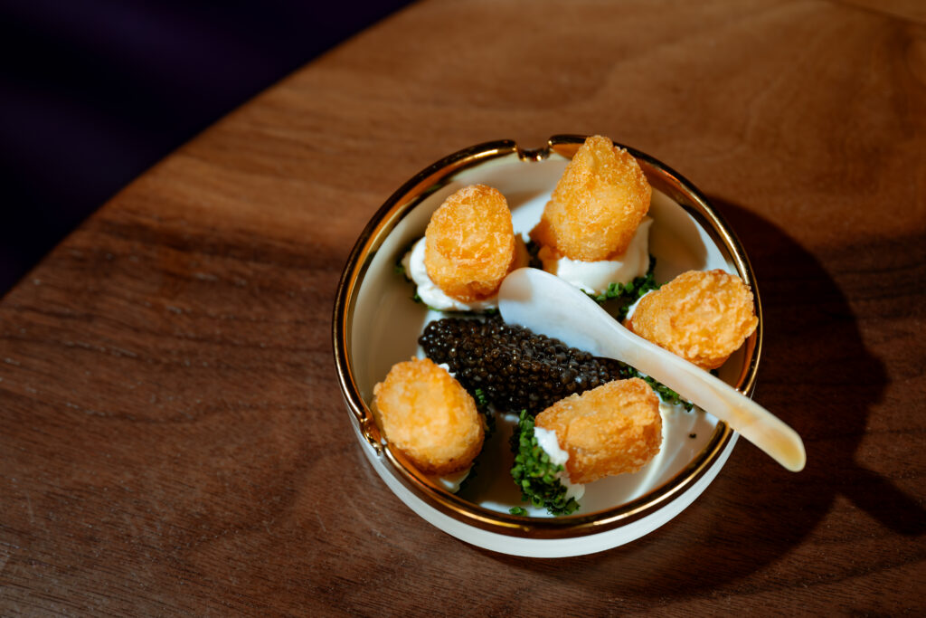 Caviar and golden potato bites arranged in a gold-rimmed ashtray with a pearlescent spoon on a wooden table.