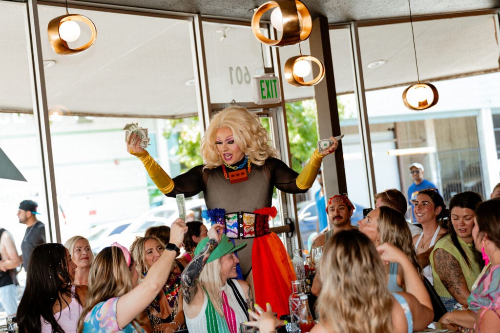 Drag performer wearing a blonde wig and colorful skirt holding dollar bills as seated brunch guests cheer at Champagne Tiger.