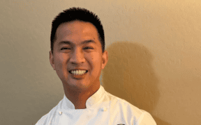 Chef Allen Rubio smiling in a white chef’s coat with his hands clasped.