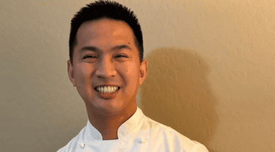 Chef Allen Rubio smiling in a white chef’s coat with his hands clasped.