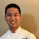 Chef Allen Rubio smiling in a white chef’s coat with his hands clasped.