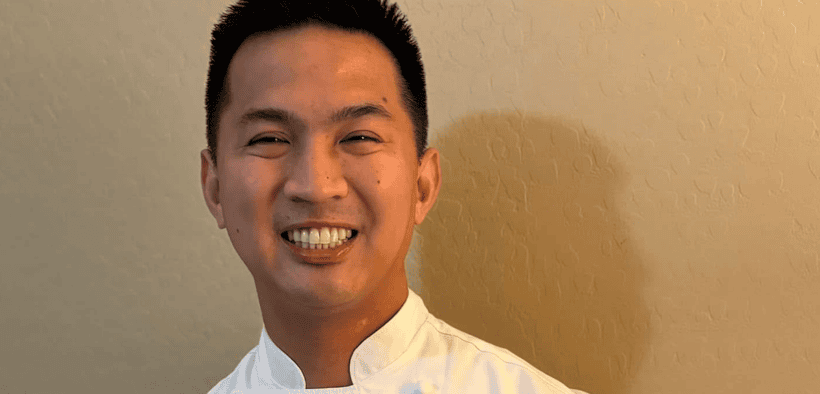 Chef Allen Rubio smiling in a white chef’s coat with his hands clasped.