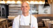 Chef Cory Oppold wearing a white chef’s coat and gray apron, standing with his arms crossed inside COURSE in Scottsdale.