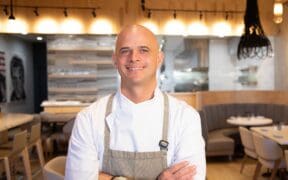 Chef Cory Oppold wearing a white chef’s coat and gray apron, standing with his arms crossed inside COURSE in Scottsdale.