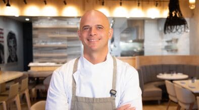 Chef Cory Oppold wearing a white chef’s coat and gray apron, standing with his arms crossed inside COURSE in Scottsdale.