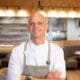 Chef Cory Oppold wearing a white chef’s coat and gray apron, standing with his arms crossed inside COURSE in Scottsdale.