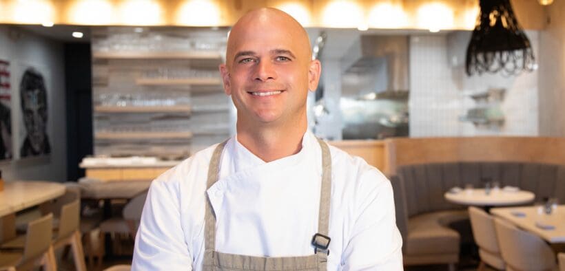 Chef Cory Oppold wearing a white chef’s coat and gray apron, standing with his arms crossed inside COURSE in Scottsdale.