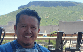Chef Jesus Silva smiling in a blue shirt and gray apron on an outdoor patio with a mountain backdrop.