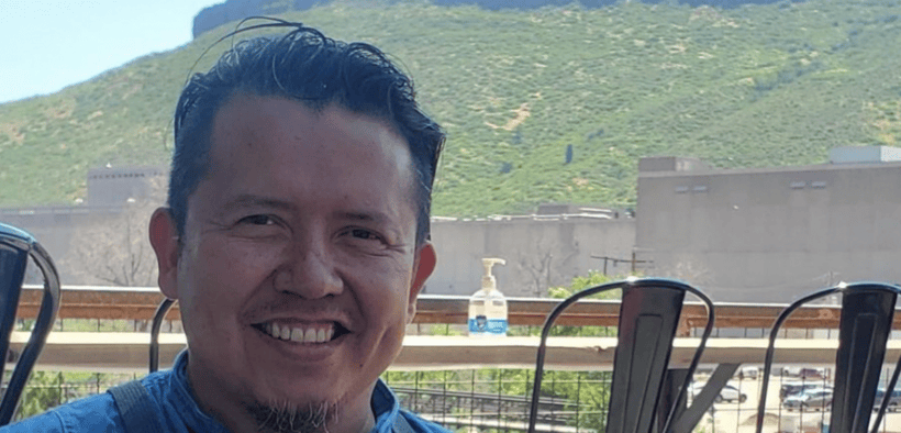 Chef Jesus Silva smiling in a blue shirt and gray apron on an outdoor patio with a mountain backdrop.