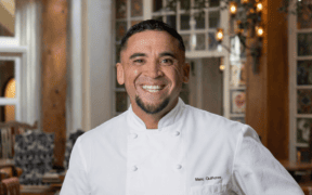 Chef Marc Quiñones smiling in a white chef’s coat inside La Plazuela in Santa Fe.