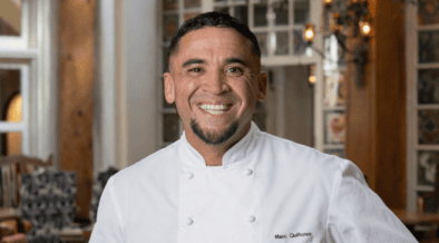 Chef Marc Quiñones smiling in a white chef’s coat inside La Plazuela in Santa Fe.