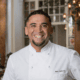 Chef Marc Quiñones smiling in a white chef’s coat inside La Plazuela in Santa Fe.