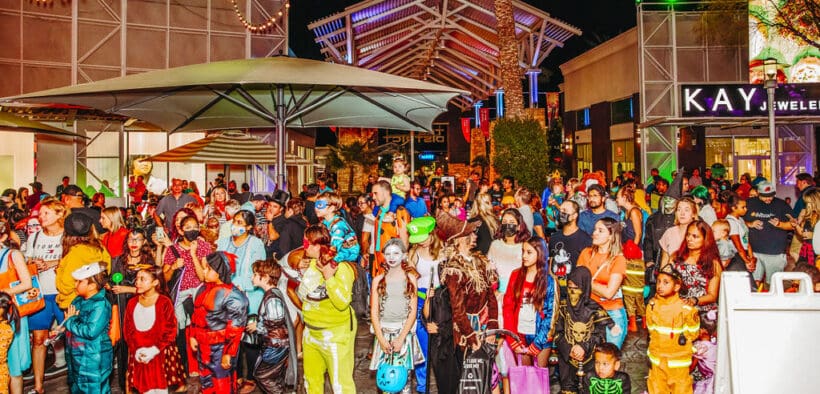 Large crowd of adults and children in Halloween costumes at Desert Ridge Marketplace during an evening celebration.