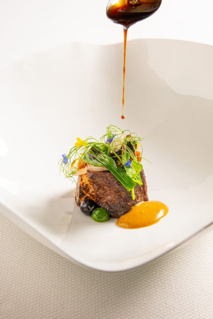 Sauce being drizzled over a composed dish topped with greens and edible flowers at Kai.