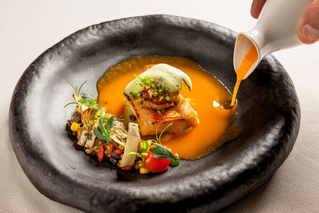 Orange sauce being poured around a composed entrée with vegetables and fresh herbs at Kai.