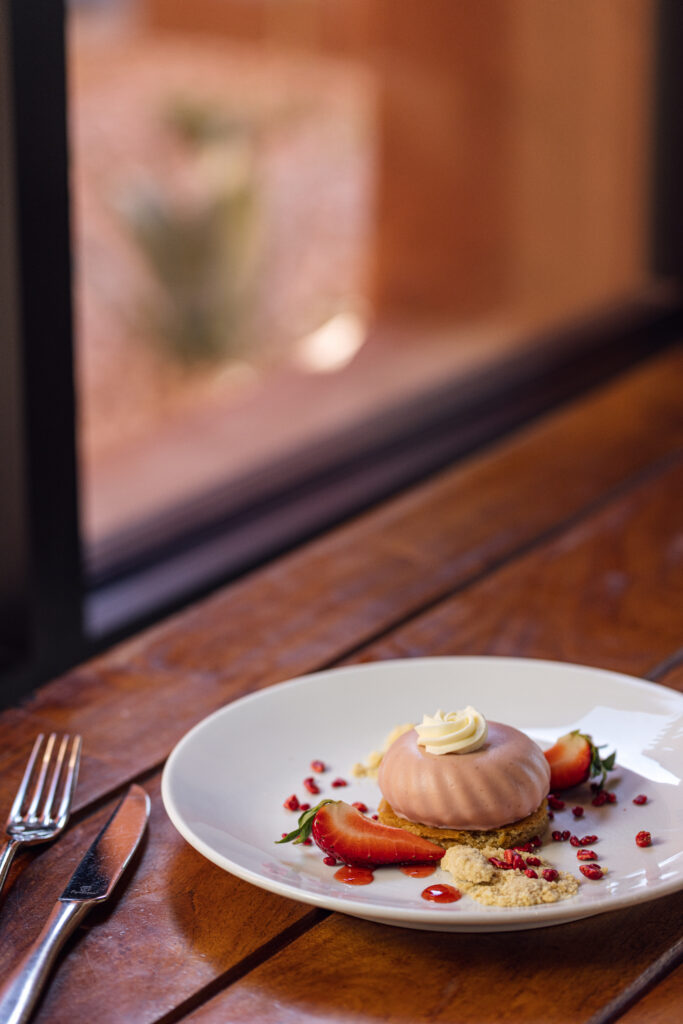 Round pink-glazed dessert topped with cream and served with strawberries and crumbs on a white plate.