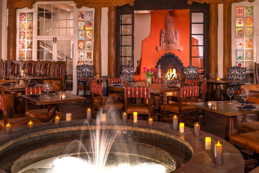 Elevated view of La Plazuela’s dining room with a central fountain, tall potted trees, wooden tables and colorful windows.