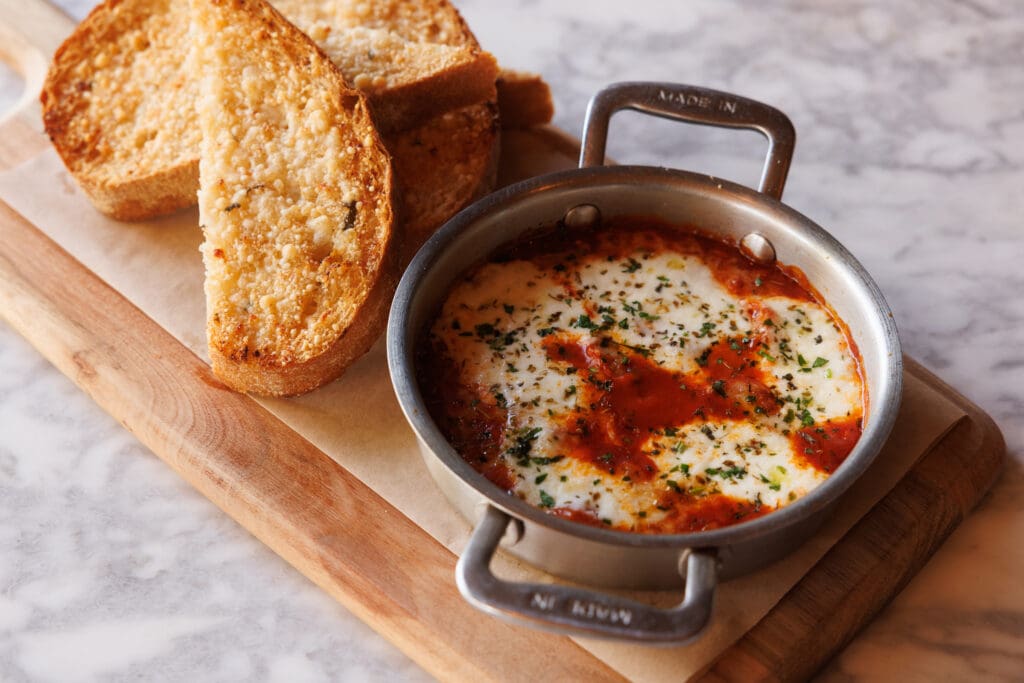 Baked burrata in red sauce topped with herbs in a small metal pan beside toasted bread.