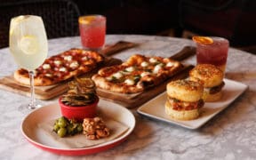 North Italia happy hour spread with flatbreads, chicken sliders, small bites and three drinks on a marble table.