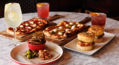 North Italia happy hour spread with flatbreads, chicken sliders, small bites and three drinks on a marble table.