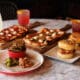 North Italia happy hour spread with flatbreads, chicken sliders, small bites and three drinks on a marble table.