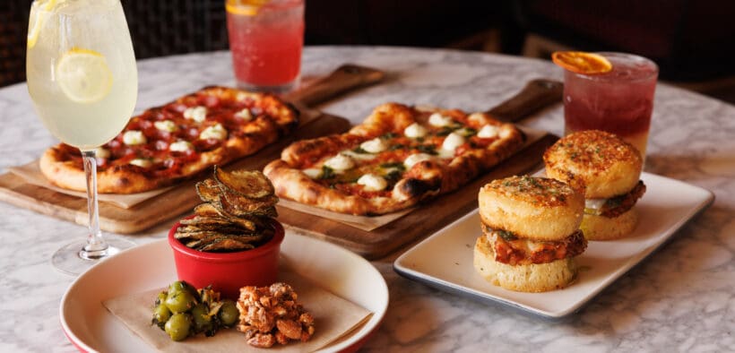 North Italia happy hour spread with flatbreads, chicken sliders, small bites and three drinks on a marble table.