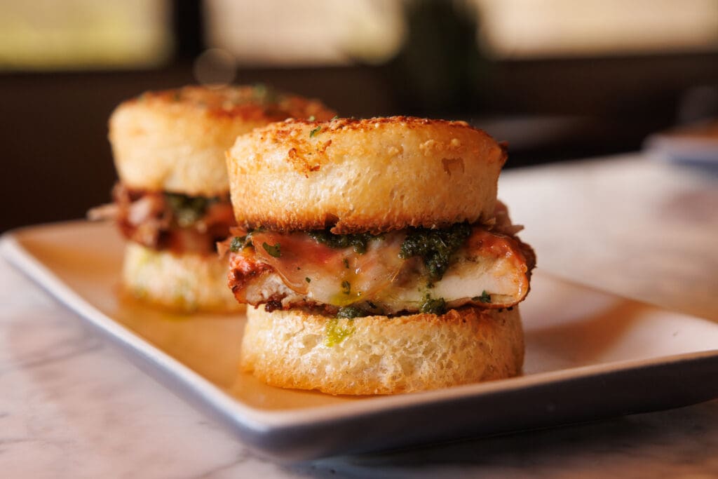 Two chicken pesto sliders with melted cheese served between golden garlic knot buns on a rectangular plate.