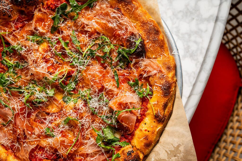 Close-up of North Italia’s Hot Honey & Smoked Prosciutto Pizza topped with prosciutto, greens and grated cheese.