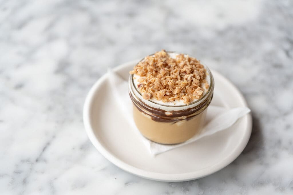 Salted caramel budino in a glass jar with a white cream layer and crumb topping on a white plate.