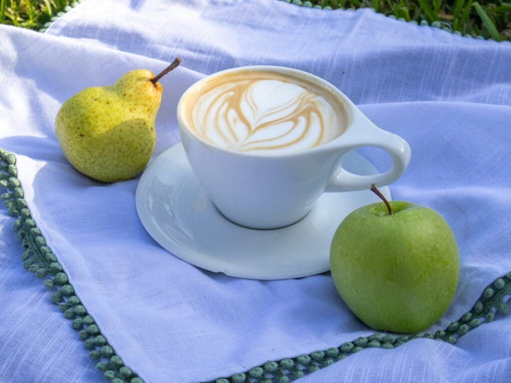 White cup of latte art surrounded by a green apple and yellow pear on a pale blue cloth outdoors.