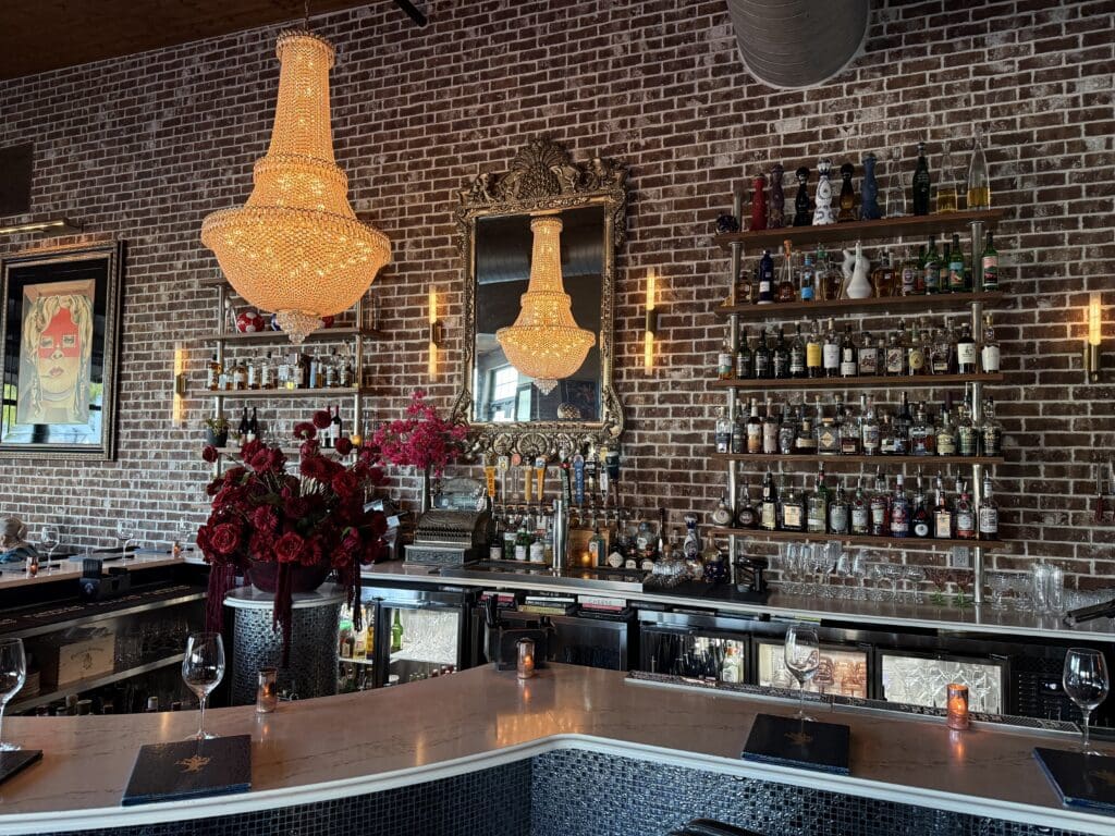 Bar at Primrose Path in College Station with crystal chandeliers, red flowers, exposed brick and shelves of spirits.
