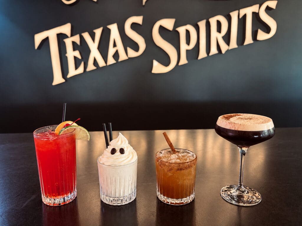 Four specialty cocktails, including a red citrus drink, a creamy cocktail and an espresso-style cocktail, displayed beneath a Texas Spirits sign at Rickhouse.
