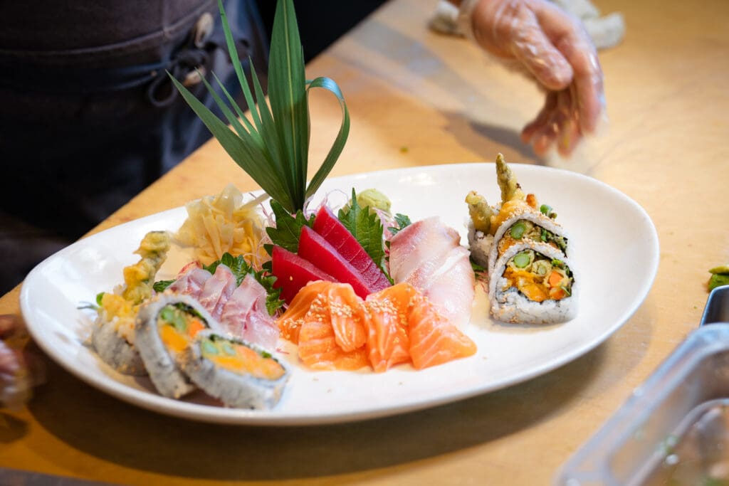 Sliced sashimi and sushi rolls arranged with ginger, wasabi and decorative green leaves on a white platter.